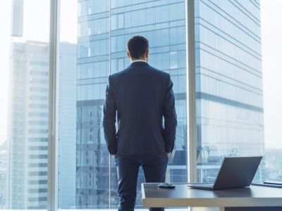 Un uomo d'affari di spalle osserva grattacieli aziendali contemporanei dalla vetrata del suo ufficio