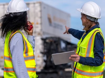 Due lavoratrici con gilet ad alta visibilità e caschi protettivi, posizionate davanti a dei container