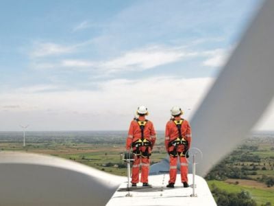 Tecnici in tuta arancione con imbracature di sicurezza sul motore di una turbina eolica, con vista panoramica su un parco eolico in campagna