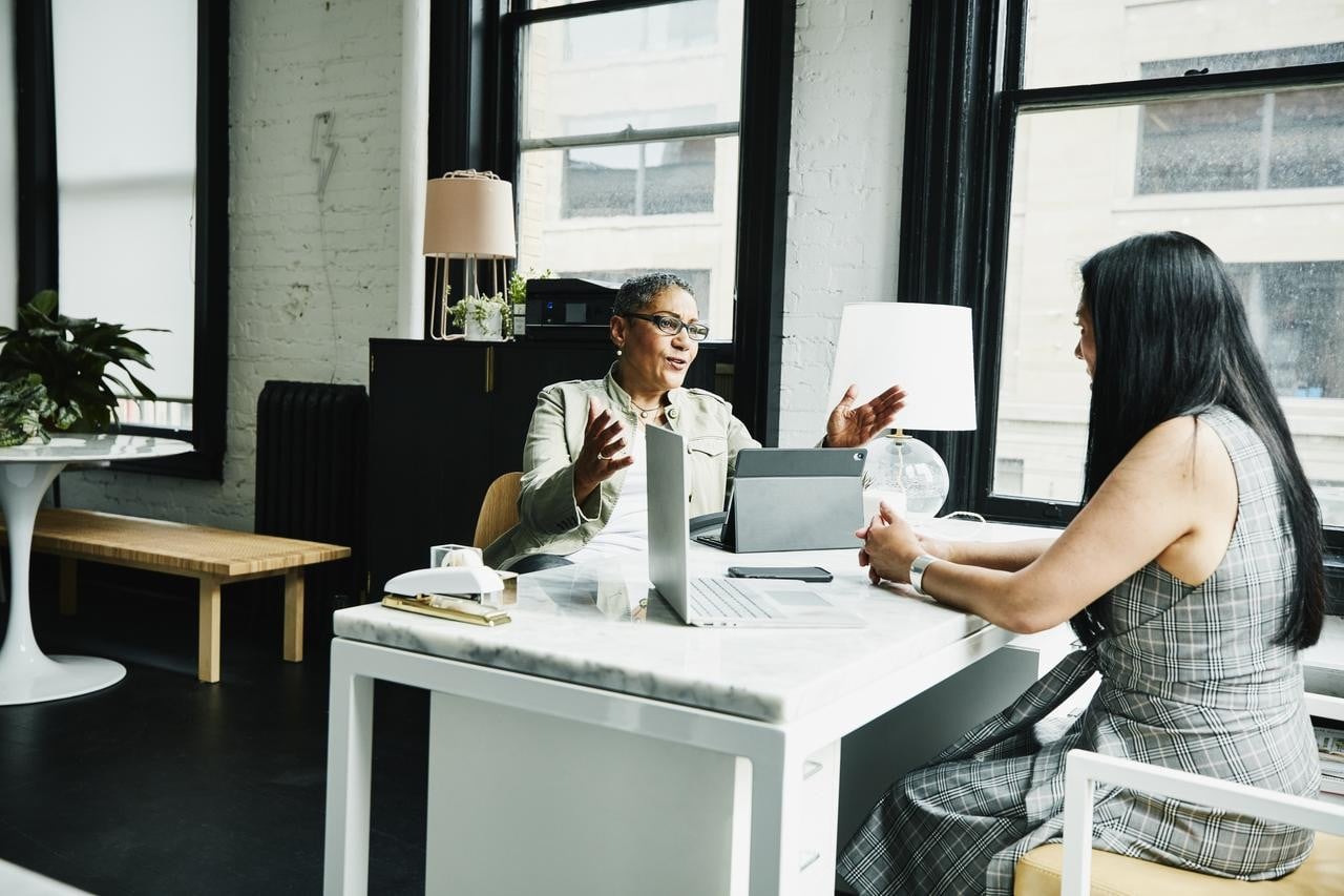two women sitting at a white desk one with a ipad and the other with her phone and computer on the table facing each other in the midst of discussion. One lady has glasses and is in the midst of talking while the other has her hands clasped together on the table listening.