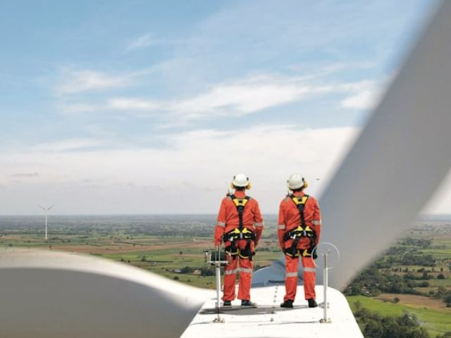 Tecnici in tuta arancione con imbracature di sicurezza sul motore di una turbina eolica, con vista panoramica su un parco eolico in campagna