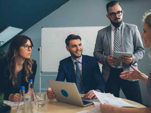 3 employees in a business meeting. Left side is women and man listening to conversation. Right side is a man standing and holding a tablet.  