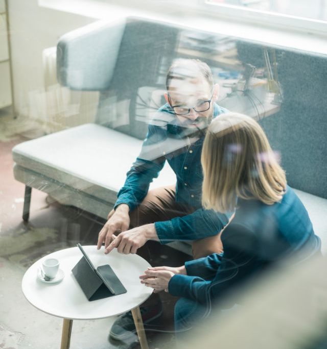 man and woman sitting and discussing business matters. In front of them is a desk with a tablet 