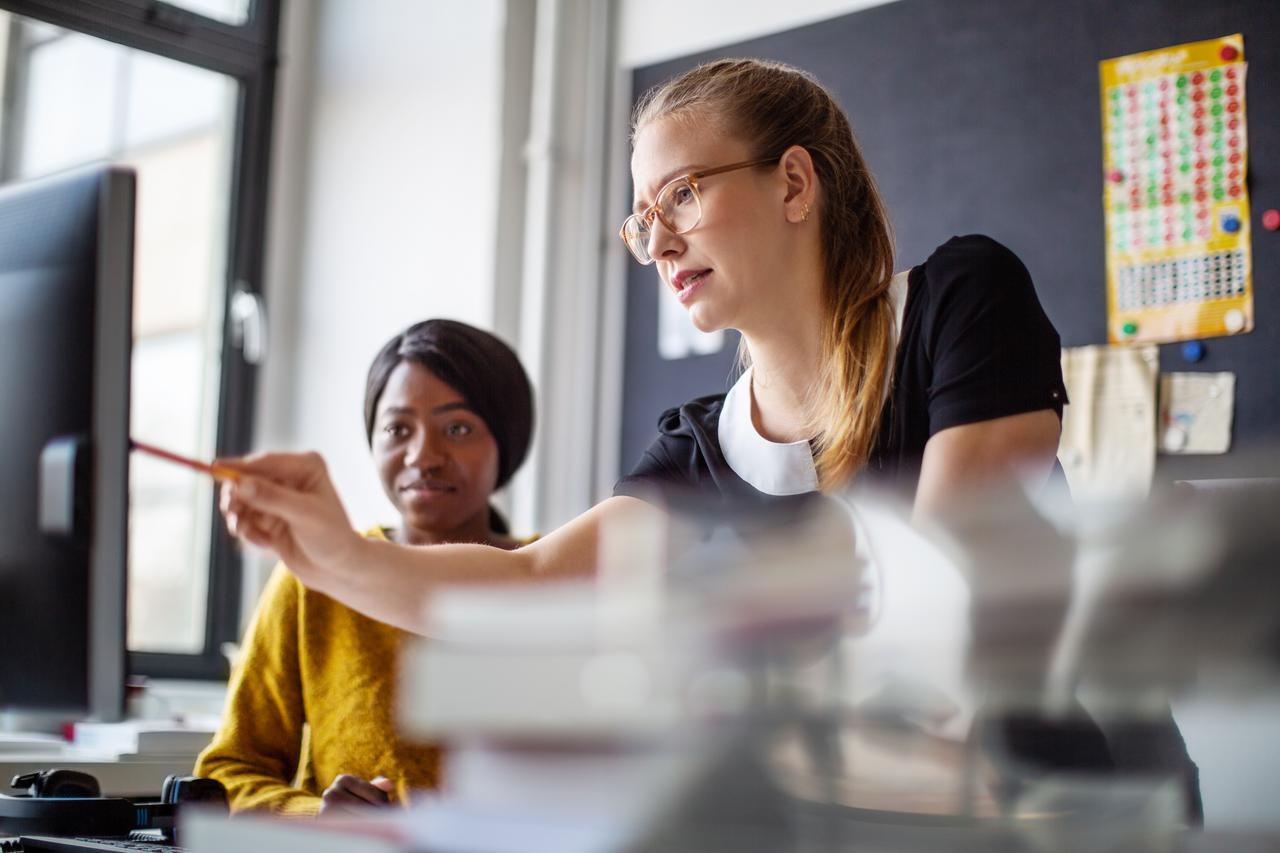 A woman points at a computer screen and explains something to another woman.