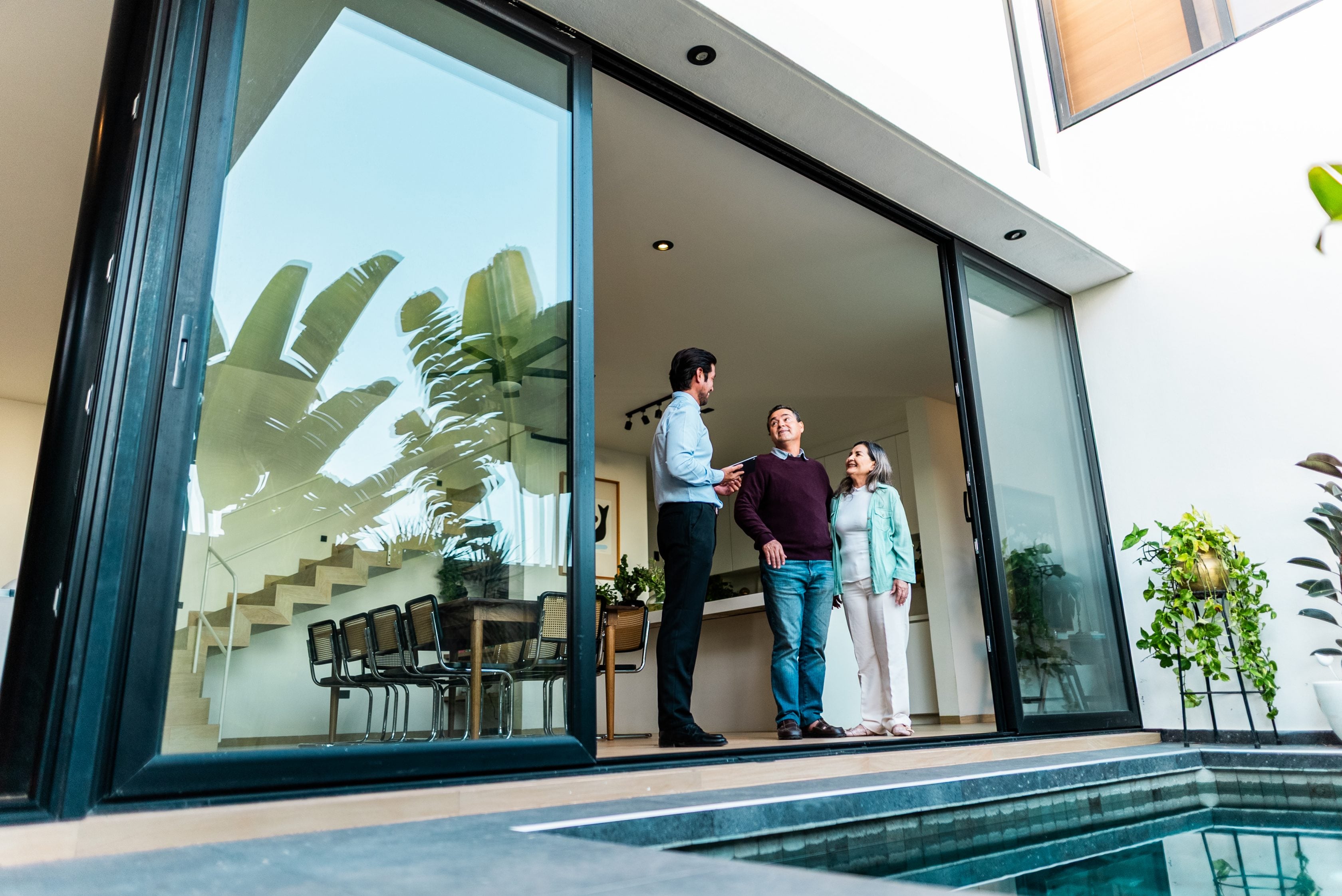 realtor and older couple standing inside of home looking out into backyard with a pool in front of them