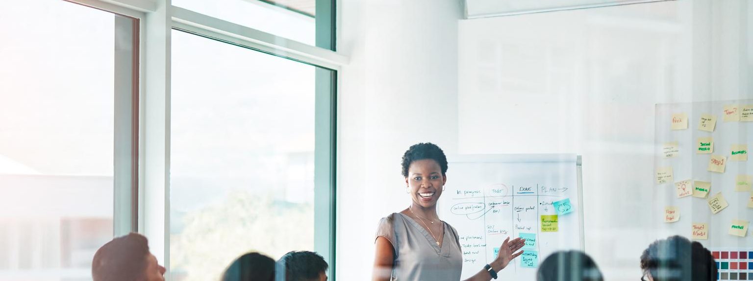 woman standing in fron of white board while room full of colleagues look at her