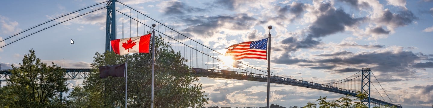 Canadian and US flag side by side with body of water and bridge in background