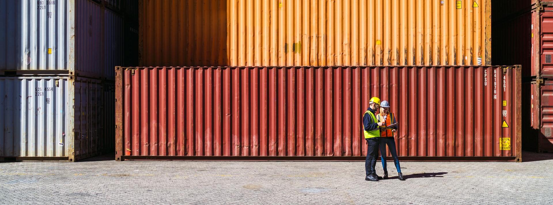 one male and one female construction worker looking at clipboard together with shipping containers in background