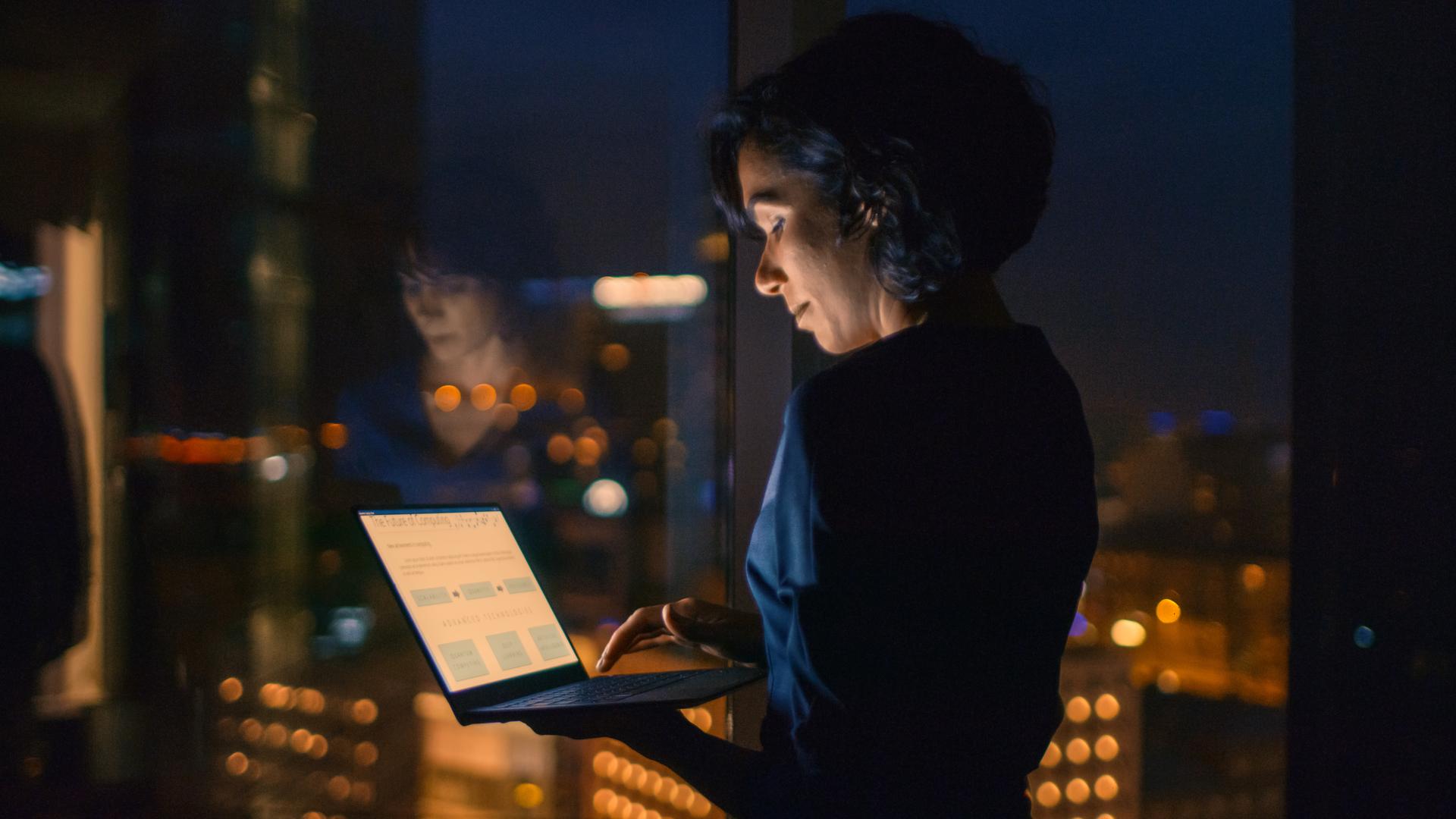 woman standing in hotel room looking at laptop with the city skyline in background at night time