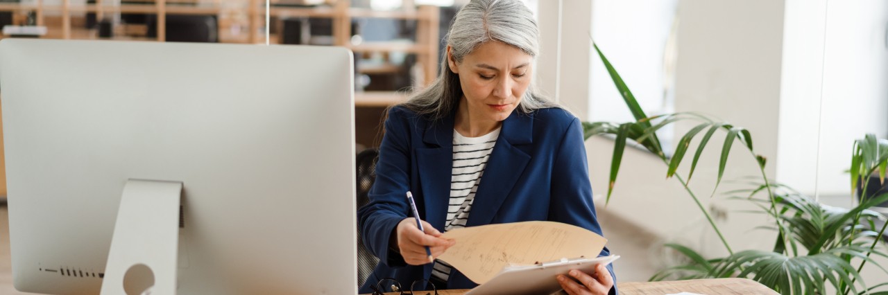 woman flipping through a folder with a pencil in her hand with a monitor in front of her. She seems to be checking through the information in the folder with what is on the monitor.