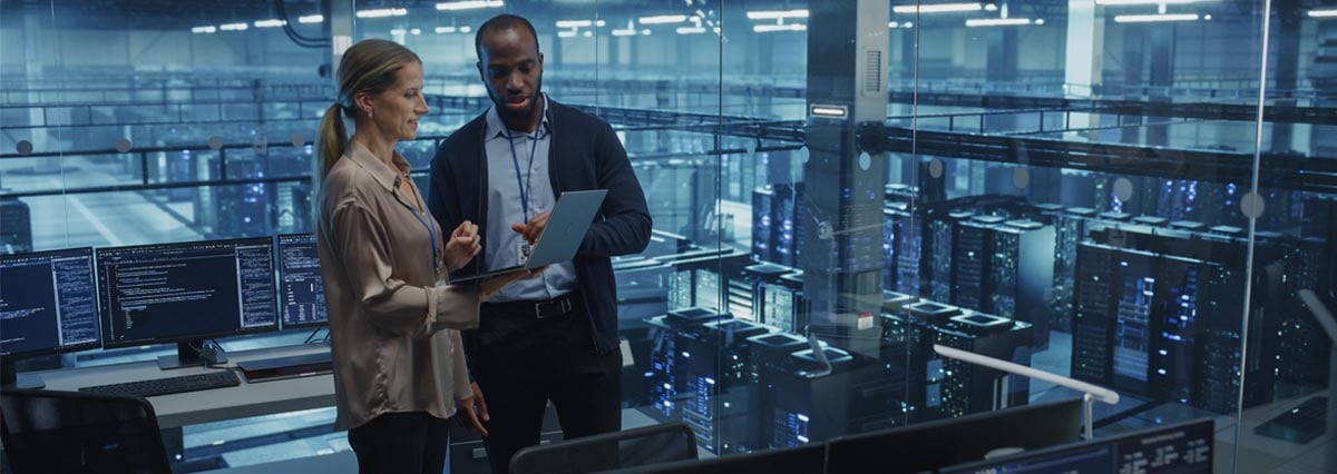 Male Cloud Architect and Female Database Engineer Review Data Storage Protocols on a Shared Laptop in a Data Center. Team Strategizing Ways to Improve Service for Enterprise Clients