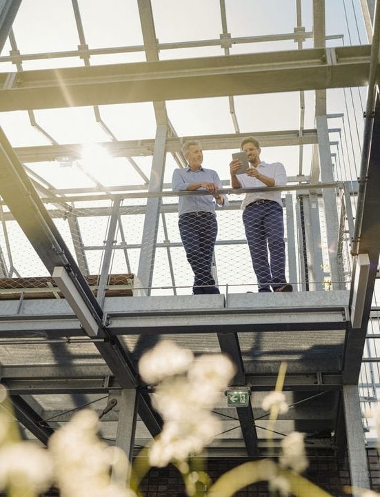 Deux hommes se tiennent sur une structure métallique en extérieur, discutant et regardant une tablette, tandis que la lumière du soleil illumine la scène en arrière-plan.