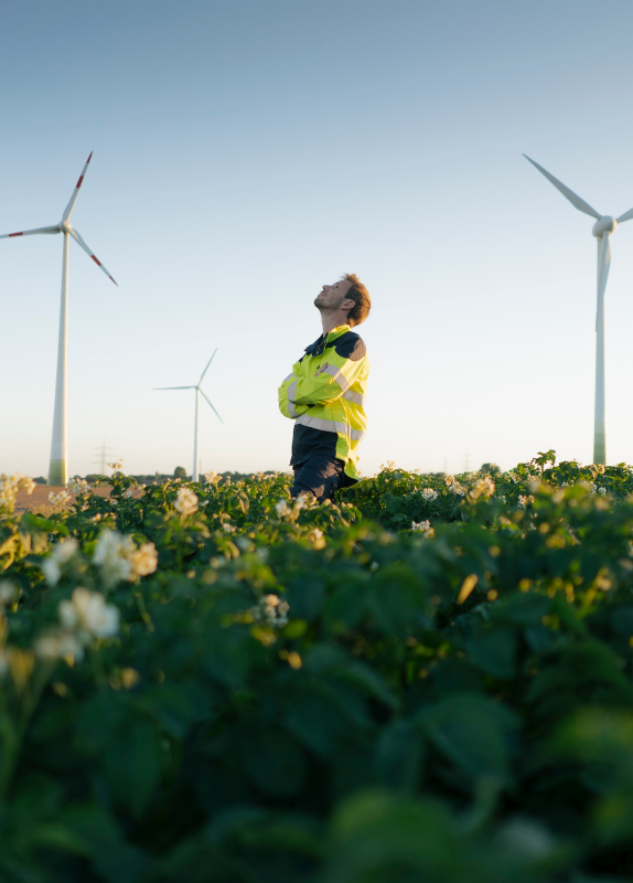 This image shows a man in a reflective safety jacket standing in a field of plants, looking up towards the sky.