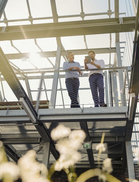 Deux hommes se tiennent sur une structure métallique en extérieur, discutant et regardant une tablette, tandis que la lumière du soleil illumine la scène en arrière-plan.