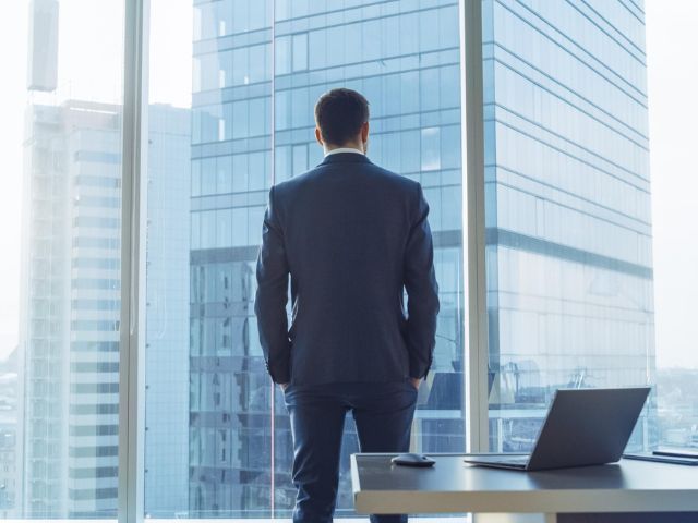 Un uomo d'affari di spalle osserva grattacieli aziendali contemporanei dalla vetrata del suo ufficio