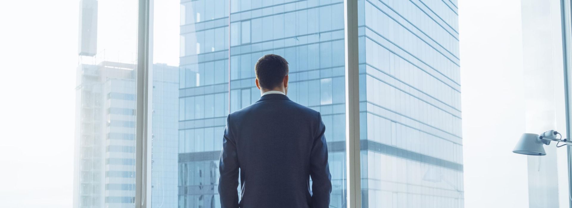 Un uomo d'affari di spalle osserva grattacieli aziendali contemporanei dalla vetrata del suo ufficio
