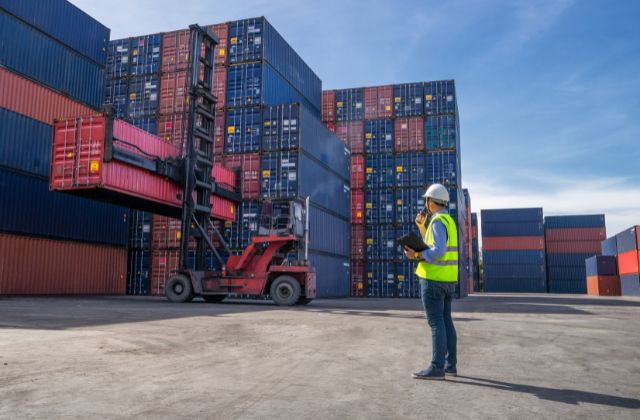 Un ingegnere dirige le operazioni logistiche in un deposito di container, indossando un casco di sicurezza e un giubbotto riflettente