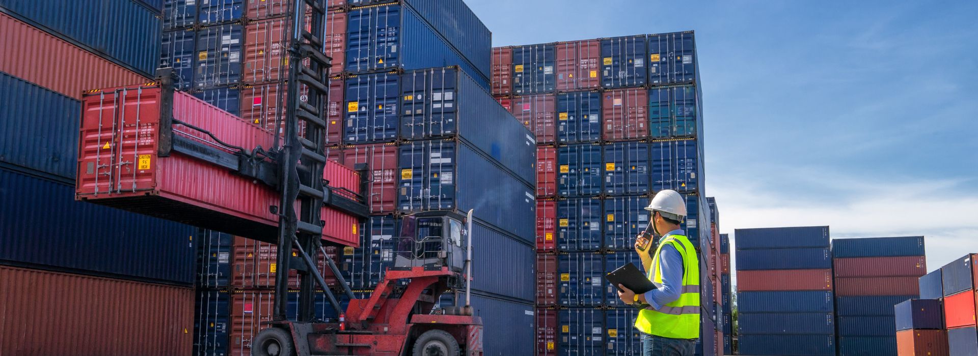 Un ingegnere dirige le operazioni logistiche in un deposito di container, indossando un casco di sicurezza e un giubbotto riflettente