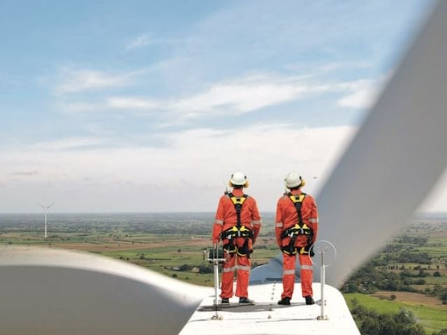 Tecnici in tuta arancione con imbracature di sicurezza sul motore di una turbina eolica, con vista panoramica su un parco eolico in campagna