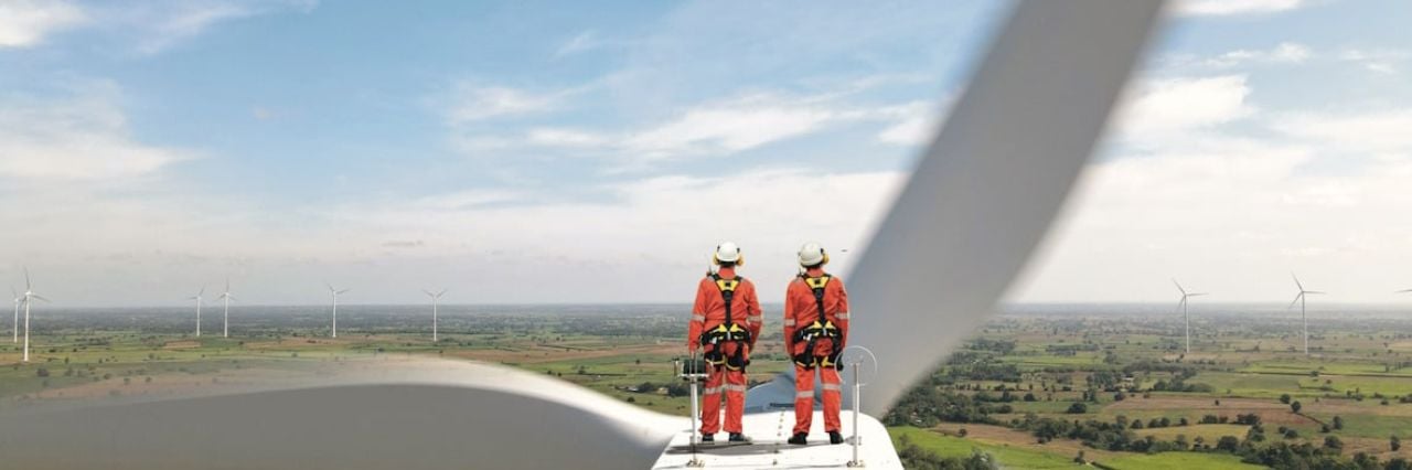 Tecnici in tuta arancione con imbracature di sicurezza sul motore di una turbina eolica, con vista panoramica su un parco eolico in campagna