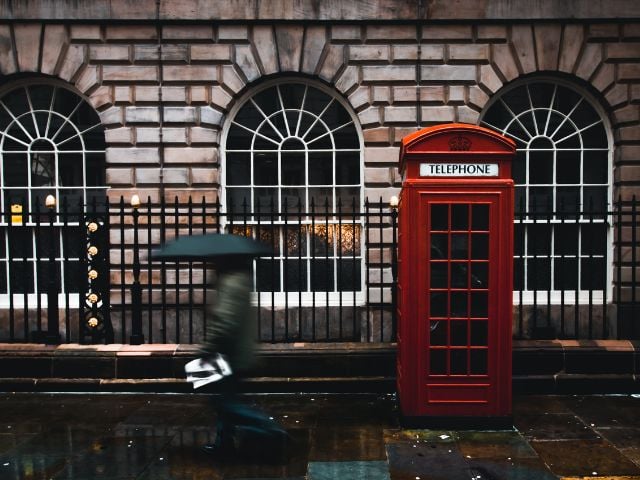 Una persona cammina con un ombrello aperto in una strada di Londra dove è presente una cabina telefonica rossa