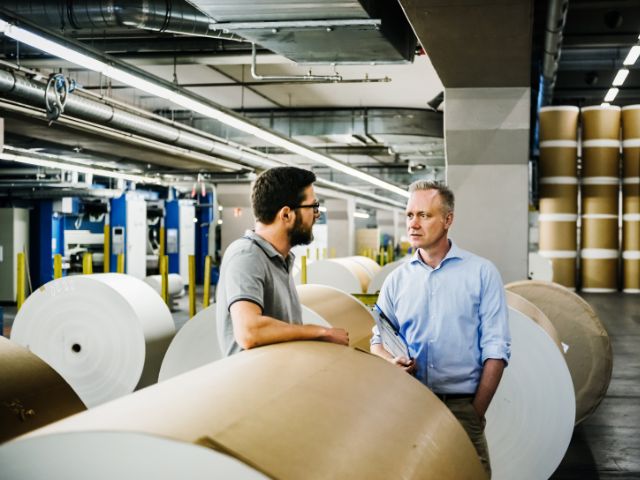 Due lavoratori in un magazzino discutono circondati da grandi rotoli di carta