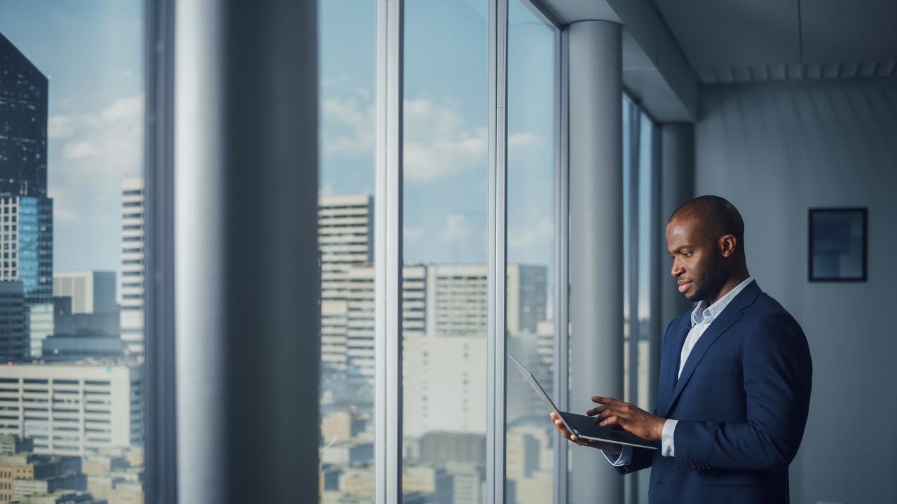 Man in een corporate office gebouw die op een laptop kijkt voor een groot raam