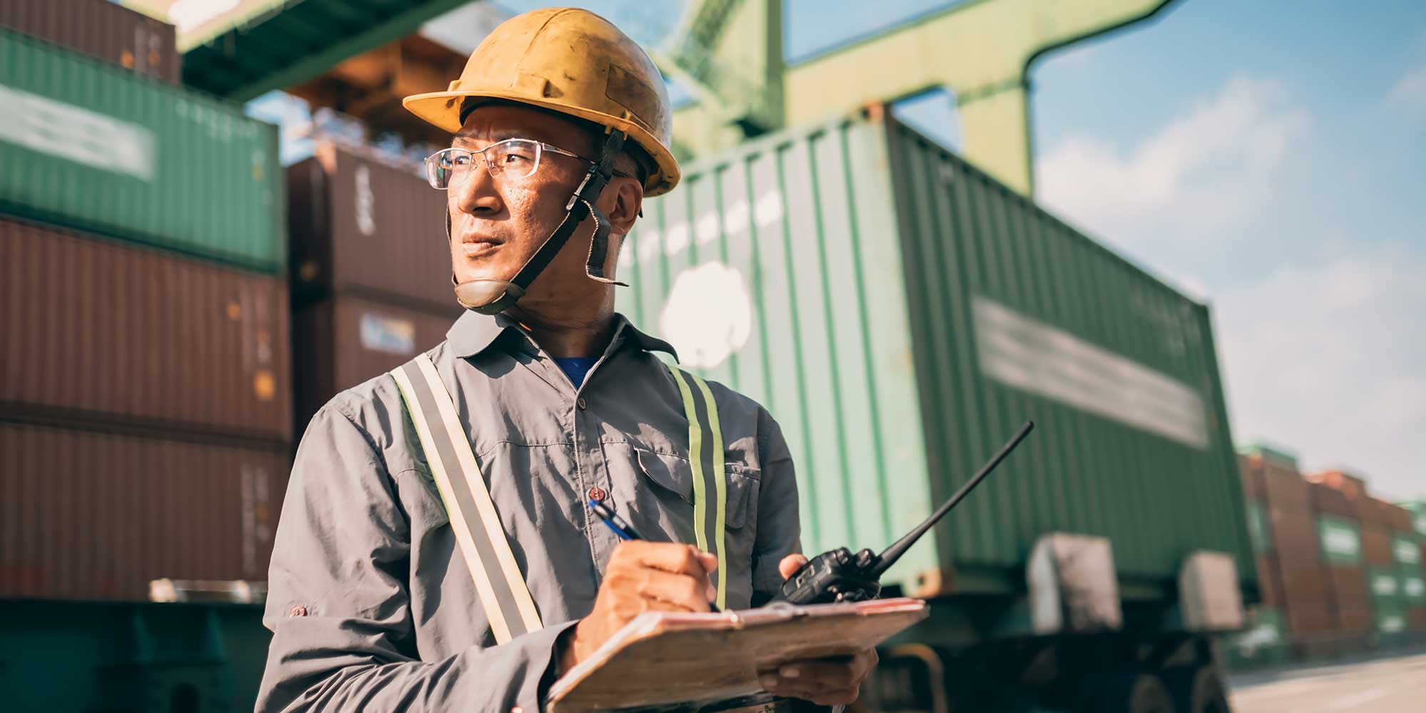 Mężczna w kasku na głowie doglada pracy w porcie przeładunkowym z kontenerami