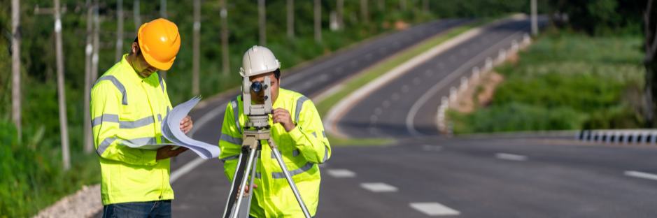 Two surveyor engineers wearing safety gear, working on a road construction site, with one using surveying equipment and the other reviewing blueprints near a newly paved road.